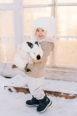 Vertical portrait of happy little child boy in warm clothes hugging white funny rabbit standing by farm house on sunny winter day, looking at camera. Concept of excursion to eco-farm, life in village.