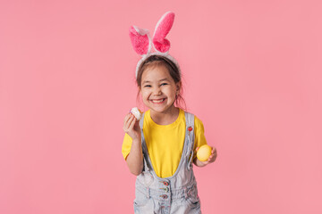 Little girl wearing bunny ears on her head and eating Easter eggs in front of a pink background.