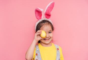 Little girl wearing bunny ears on her head holding a yellow Easter egg in her hand against a pink...
