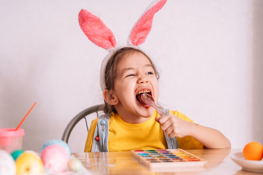 A little girl wearing bunny ears on her head eats chocolate from a spoon at a wooden table on which there are painted Easter eggs.