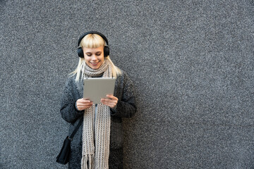 Happy hippie girl browsing a tablet and listening to the music with headphones leaning on a wall in...