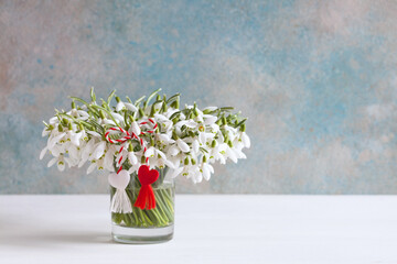 Bouquet of snowdrops in a glass on a table, a red and white symbol of a martenitsa and a heart on the background of a colorful wall. Postcard for the holiday of March 1. - 724849749