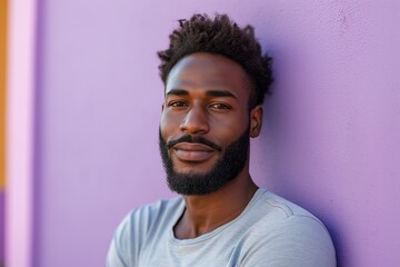 A good-looking black man with a beard and short black hair in a light-colored shirt is leaning against a purple wall