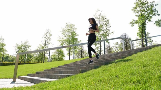 A slender young woman in black sweatpants and a tank top runs down the stairs in a modern city park. Outdoor training in summer. Female runner. Slow motion. The image is stabilized.