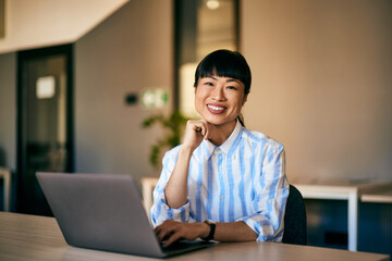 Portrait of an Asian businesswoman, smiling for the camera while working over the laptop.