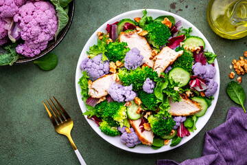 Healthy salad from purple cauliflower, broccoli, chicken breast, fresh cucumbers, onion, radicchio, spinach with walnuts, green table background, top view