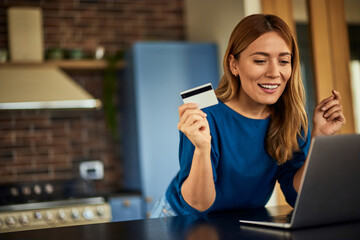 A smiling blonde woman holding a credit card, using a laptop, at home.