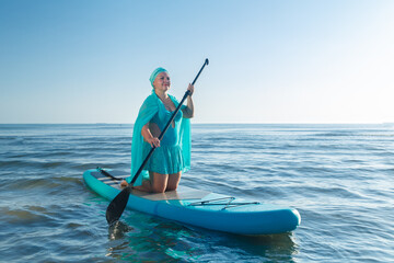 A Jewish woman in a turquoise swimsuit with a skirt and a scarf on her head kneeling on a SUP board near the sea.