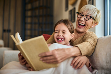 Grandma and grandchild have a great time, looking funny, and laughing while reading a book. - obrazy, fototapety, plakaty