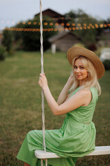 A Caucasian blonde woman in a flowing green dress enjoys a peaceful moment on a white swing in a lush garden. The concept of recreation, leisure and rural life outside the city.