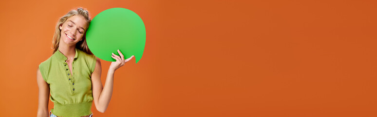 jolly teenage girl holding speech bubble and posing with closed eyes on orange backdrop, banner