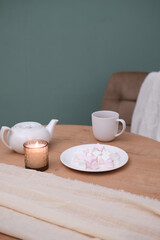 Fototapeta na wymiar Home kitchen interior - white teapot with hot tea, cup of tea, plate with marshmellow, napkin, beige chair on green background.
