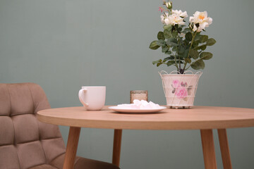 Home kitchen interior - cup of tea, plate with marshmellow, burning candle, green plant in pot, beige chair on green background.