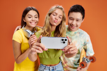 focus on smartphone in hands of three jolly blurred diverse friends taking selfie on orange backdrop