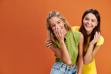 joyous adorable adolescent girls hugging and smiling at camera on orange backdrop, friendship day