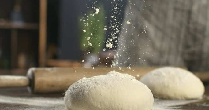Super slow motion macro of professional artisan baker chef sprinkles flour on raw loaf of dough while making homemade bread, pasta or pizza on rustic wooden table in traditional bakery kitchen.