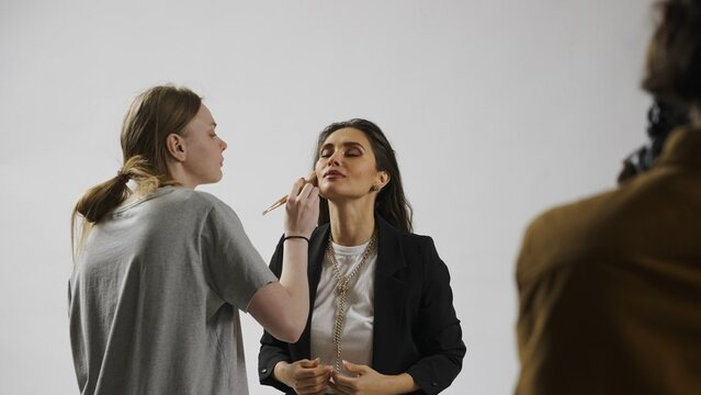 Backstage of model and professional team in the studio. Close up of model and assistant stylist fixing makeup on set, photographer taking shots.