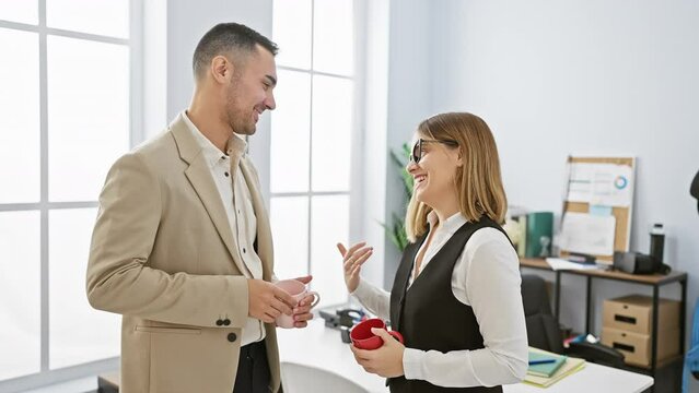 A man and woman converse happily in a bright office, both holding mugs, portraying a professional and amicable team atmosphere.