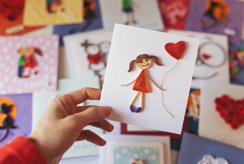 Hand holding quilling card with stick figure girl with heart balloon. woman making greeting cards. Hand made of paper quilling technique. Handicraft at home. Hobby, home office.