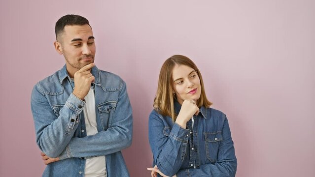Beautiful couple in denim shirts, hands on chins, standing over a pink isolated background. thoughtful faces filled with doubt pondering over a puzzling question; a picture of deep thinking.