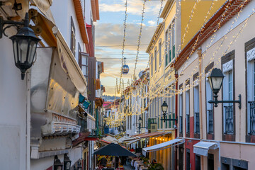 A cable car gondola to the town of Monte passes over the famous Rua de Santa Maria narrow street of cafes, colorful doors and shops in the historic medieval old town of Funchal, Madeira Portugal. - obrazy, fototapety, plakaty