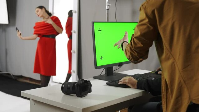 Model and production team in the studio. Photographer looks at chroma key green screen computer, model taking selfie pics.