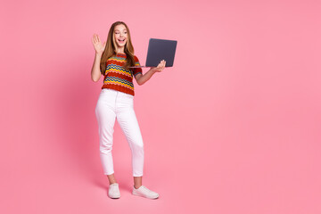 Full length photo of friendly positive girl arm palm waving hi speak video call netbook empty space isolated on pink color background