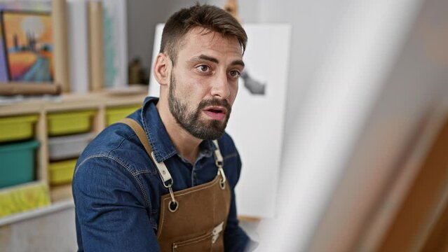 Relaxed young hispanic man, a portrait of handsome bearded artist, focused and concentrating on drawing with paintbrushes at art studio, perfecting canvas masterpiece