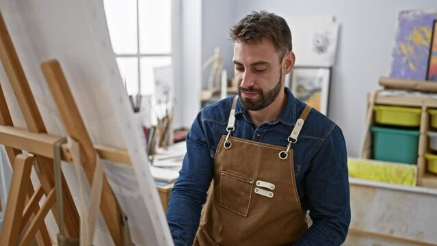 Relaxed young hispanic man, a portrait of handsome bearded artist, focused and concentrating on drawing with paintbrushes at art studio, perfecting canvas masterpiece