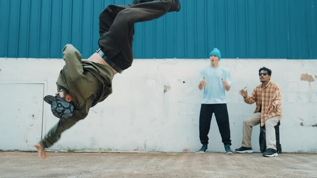 Skilled hipster doing cart wheel while multicultural friends encourage him. Hip-hop dancer moving foot step to music surrounded by group of hipster at wall near street. Outdoor sport 2024. hiphop.