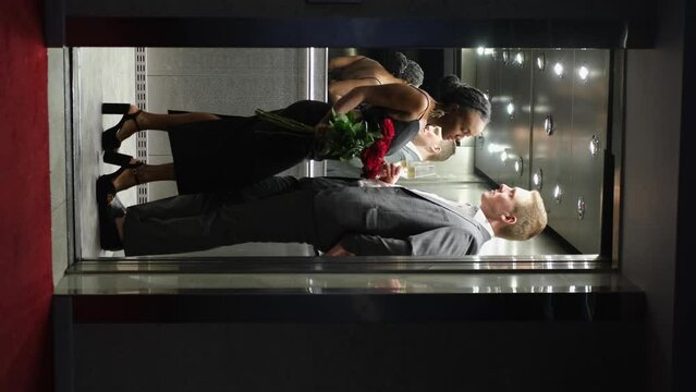 A handsome young man and a beautiful woman look at each other and smile while standing inside an elevator. Couple of lovers with glasses of champagne and a bouquet of roses in an elevator - Powered by Adobe