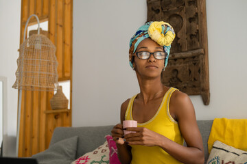 Portrait of pensive young black woman wearing glasses and headwrap while drinking a cup of coffee at home. - 724960761