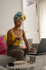 Cheerful black woman working at home with laptop while taking a cup of coffee. African style young black female model wearing headwrap. - 724960766