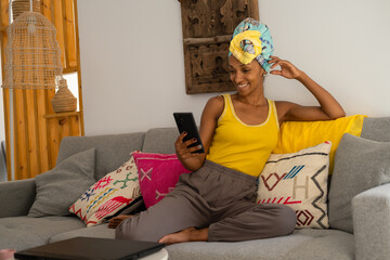 Relaxed black woman relaxing on the couch at home and using his smartphone.. African style young black female model wearing headwrap. - 724960769