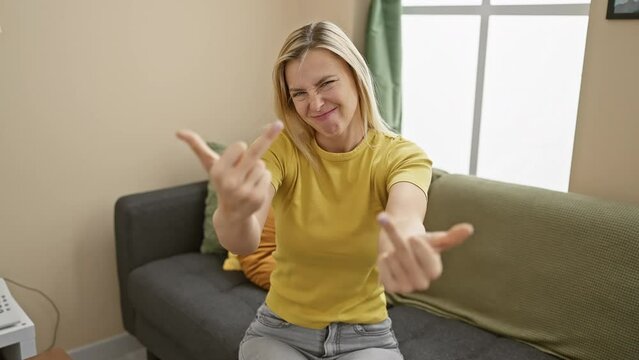 Angry blonde woman throwing 'fuck you' sign at home, showcasing her brash humor in a rude gesture of provocation. her t shirt, middle finger and gritty expression all scream 'i'm confident!'
