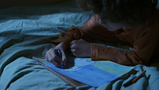Side handheld footage of little boy drawing puppy with wax crayons lying sleepless in bed at bedtime