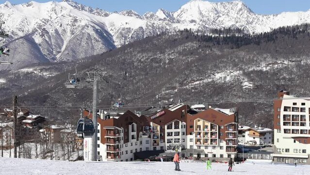 Snowboarders and skiers on slope near hotel buildings on sunny day, 4K.