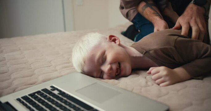 Close-up shot of a small albino boy with white hair in a brown sweater lying on the bed while his father tickles him lying near the laptop. Happy fun time for parents and children during vacation and