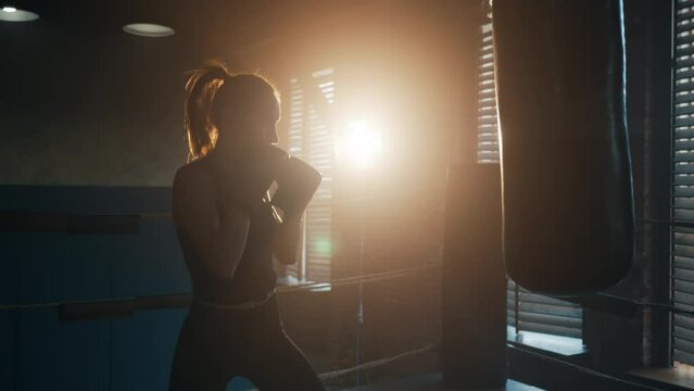 Silhouette of young tired exhausted sportswoman boxer loaning head and hands to punching bag in gym, side view. Sweaty girl is resting after hard training. Workout, box, professional sport concept.