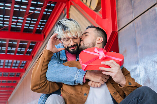 Young adult couple gay sitting outside celebrating valentine's day, his boyfriend is reciving a gift with heart shape and he is kissing him with emotion and affection for the surprise.