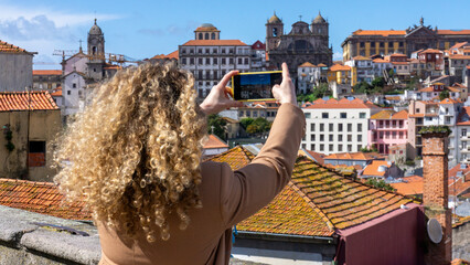 Blonde tourist woman with curly hair takes a photo with the mobile phone in the city of Porto, Portugal - obrazy, fototapety, plakaty