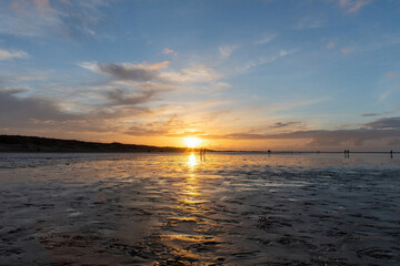 Low angle view of patch of sand along the beach at low tide along the coastline of Texel Island, the Netherlands during sunset with mix of dramatic clouds and clear skies; sun reflected in water pools