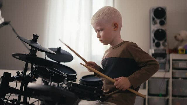 Shot from the side of a small albino boy with white hair and blue eyes playing and tuning his electric musical drum kit using special wooden sticks during his rest and day off in his room