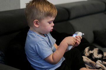 Toddler boy holding and looking at a digital thermometer, sitting on a couch, blurry background