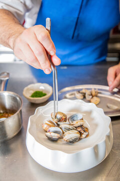 Chef decora plato de almejas a la marinera con cebollino y salsa de vino blanco albari&ntilde;o sobre un plato blanco en la barra de un restaurante