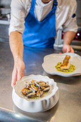 Chef decora plato de almejas a la marinera con cebollino y salsa de vino blanco albariño sobre un plato blanco en la barra de un restaurante