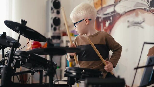 A small talented albino boy with white hair in blue round glasses plays an electronic drum musical instrument using special wooden sticks during his leisure time during the day in his room