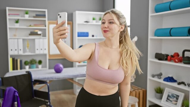 Blonde woman takes selfie in a gym with smartphone showing joy and confidence.
