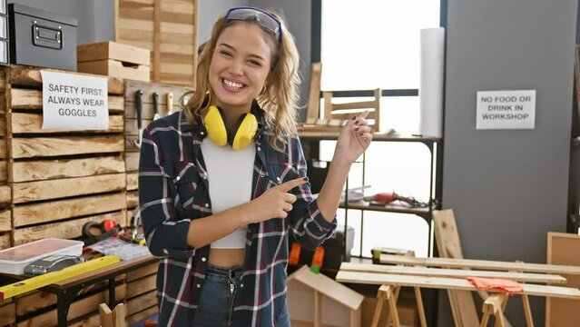 Smiling young hispanic woman showing off carpentry safety, pointing with both hands to the side while rocking headphones and security glasses