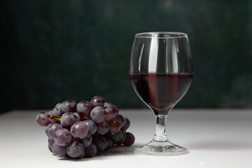Bunch of grapes with glass of wine next to them on a white table and dark background. Clean space for text.